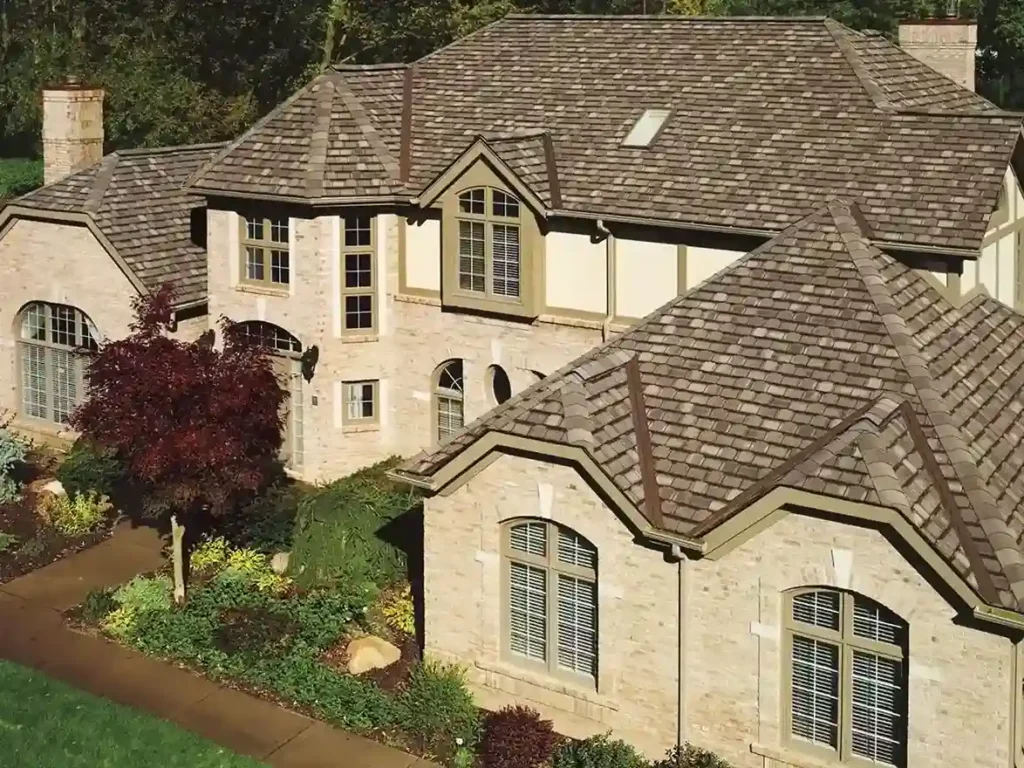 Large two-story house with a complex gabled roof installed by professional roofers, beige brick exterior, multiple windows, and a landscaped front yard with trees and shrubs.
