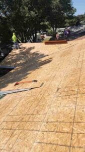 Workers stand on a roof covered with plywood sheets during a roof repair, with tools scattered around and trees visible in the background.