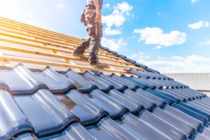 Worker standing on a sloped roof partially covered with blue ceramic tiles, applying roof waterproofing under a bright blue sky with clouds.
