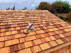 A newly installed wooden shingle roof, featuring some of the best roofing materials Fort Worth has to offer, with a hammer and nails resting on top and trees and a fence visible in the background.