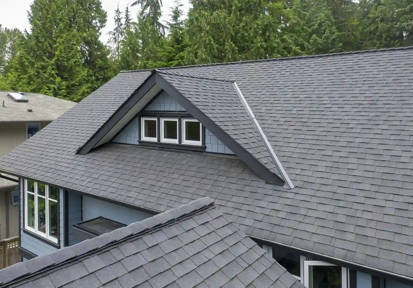 Dark gray asphalt shingle roof with a dormer and four small windows, surrounded by trees. The house exterior is blue with white trim.