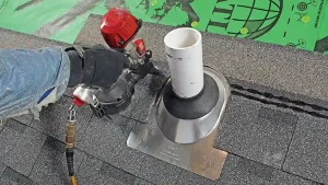 A person uses a pneumatic nail gun to secure shingles around a roof vent pipe, with underlayment and roof flashing visible—an example often seen in a comprehensive roof flashing guide.