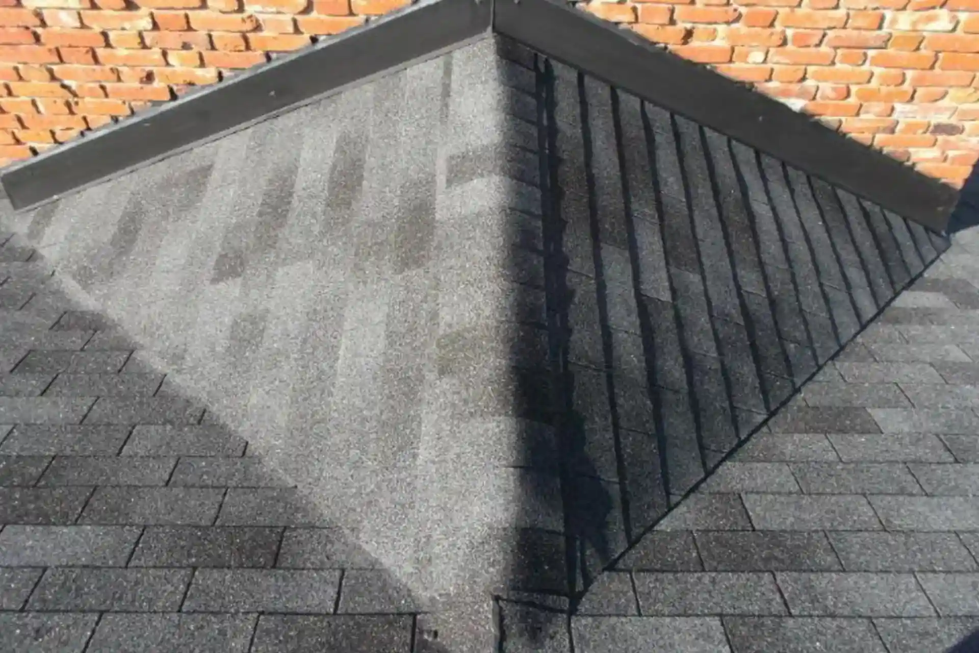 A roof corner with gray asphalt shingles, divided by a ridge; sunlight creates sharp shadow patterns on one side, while the other, featuring a roof cricket for roof leak prevention, appears uniformly lit.