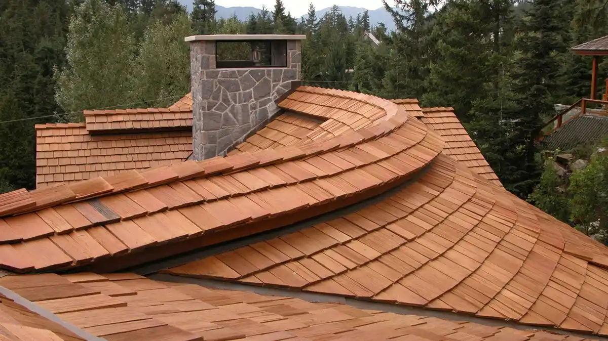 Curved cedar shake roof with a stone chimney, surrounded by trees and distant mountains in the background, highlighting the timeless appeal and natural cedar shake benefit.