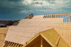 A worker installs plywood sheets on the wooden frame of a house roof under a cloudy sky, demonstrating essential roof decking techniques.