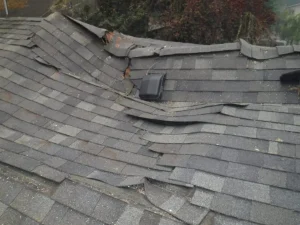 Gray asphalt shingle roof with significant buckling and visible damage, including loose and lifted shingles and a small black vent—issues that may require attention as outlined in a typical roof leak repair cost guide.