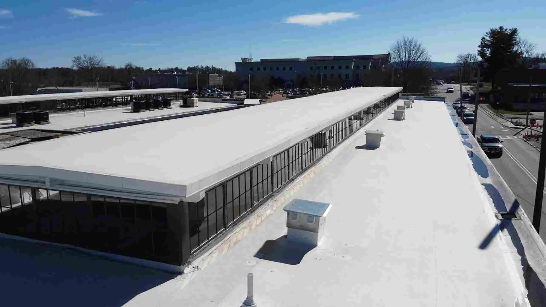 A flat white commercial roof featuring TPO roofing with HVAC units and vents, adjacent to a road and surrounded by trees and buildings under a clear blue sky—highlighting the pros and cons of TPO roofing in a practical setting.