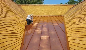 A person in a hat kneels on a large, curved cedar shake roof with smooth planks and a central copper section, outdoors on a sunny day.
