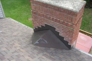 A brick chimney with step flashing and counter flashing installed at its base on a shingled roof, showcasing recommended roof flashing types from any comprehensive roof flashing guide.
