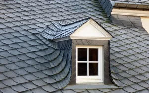 A small dormer window with white trim is set into a roof covered with overlapping slate roof tiles, arranged in a gray fish-scale pattern.