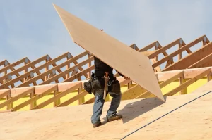 A construction worker carries a large sheet of plywood up a sloped roof while wearing a tool belt, with wooden roof trusses in the background—demonstrating techniques from the Roof Decking Guide.