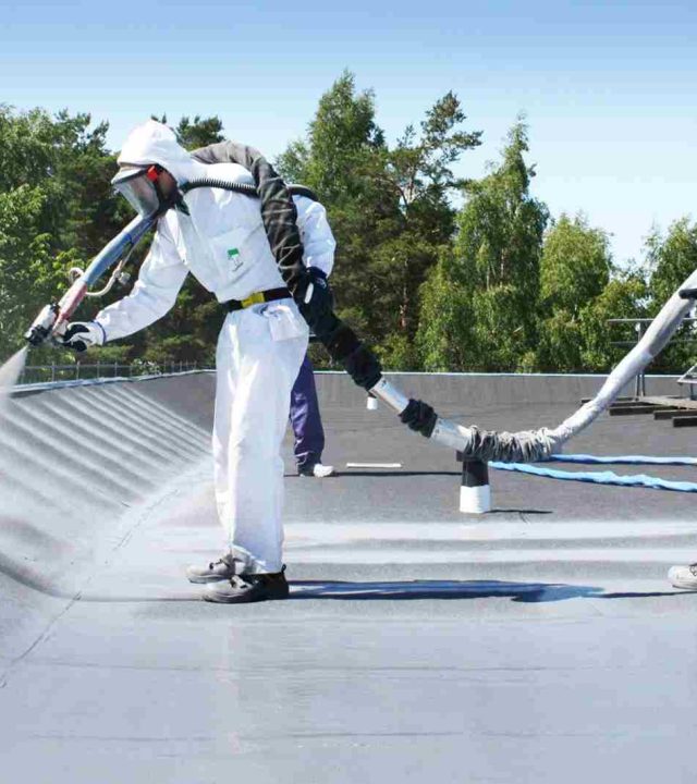ROOF WATERPROOFING 2_11zon Two workers in protective suits and masks spray roof waterproofing material onto a flat rooftop surface, with trees visible in the background.
