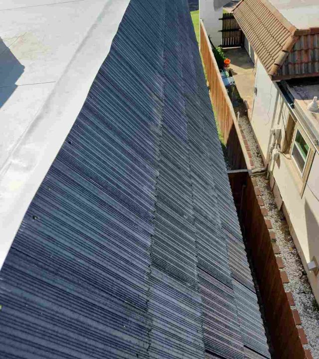 ROOF WATERPROOFING 4 A sloped roof covered with dark gray shingles, featuring roof waterproofing, is shown next to a flat white roof and a nearby street with cars and houses.