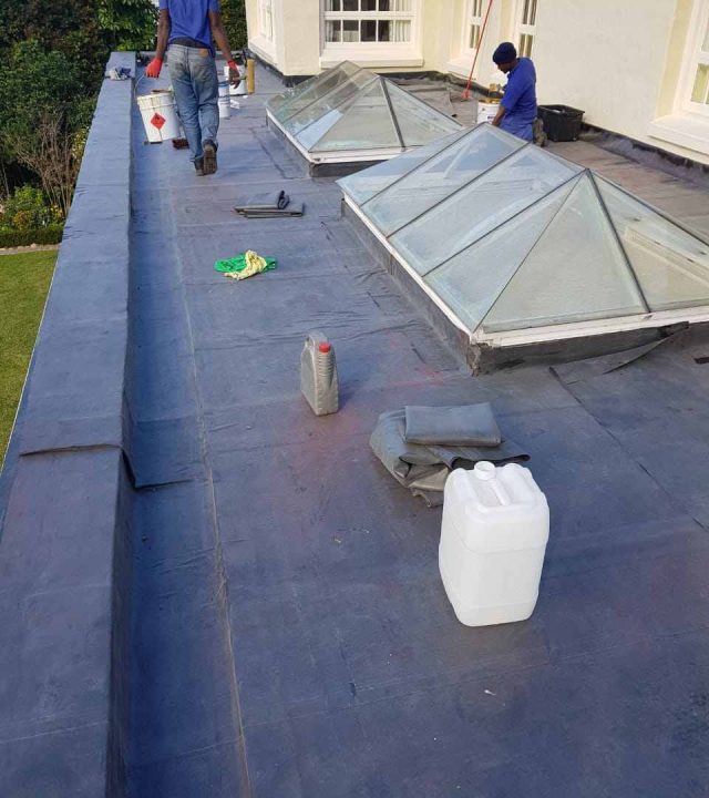 Two workers are on a flat rooftop near skylights, with various containers and tools scattered around, performing roof waterproofing and maintenance tasks.