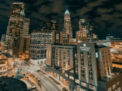 A nighttime cityscape featuring tall illuminated office buildings and the Embassy Suites hotel in the foreground under a cloudy sky, with signage from a leading roofing company North Carolina visible amid the glowing lights.