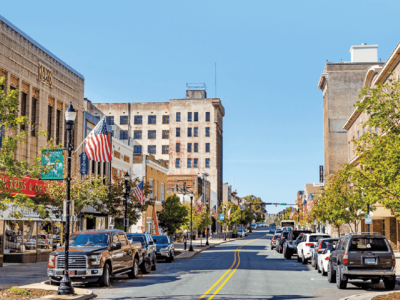 A view of a downtown street lined with parked cars, trees, storefronts, and American flags on a clear, sunny day—perfect for spotting a trusted roofing company NC as you stroll by.