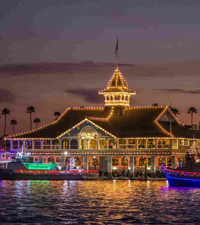 A waterfront building decorated with string lights at dusk, perfectly showcasing the craftsmanship of local roofers, with boats adorned in colorful holiday lights docked nearby on the water.